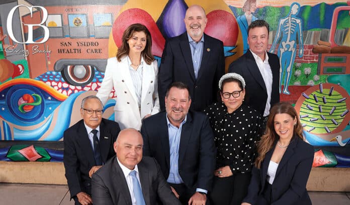 Eight professionals in business attire pose for a team photo in front of a colorful mural at San Ysidro Health Care.