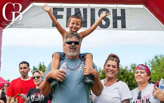 Man with a child on his shoulders crosses the finish line as supporters cheer under a banner reading FINISH.