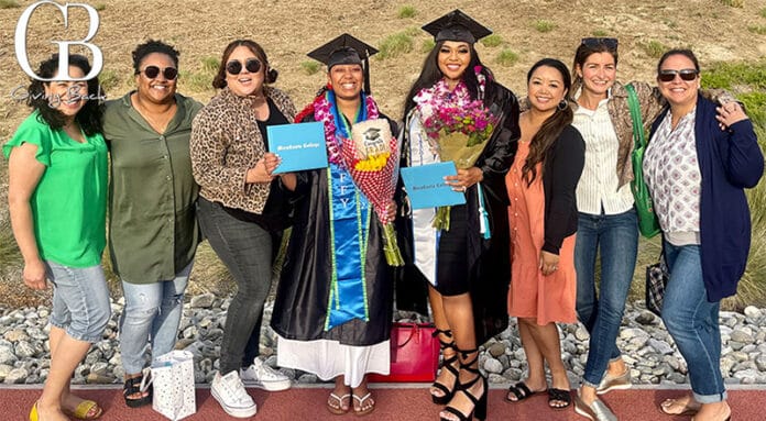 Group of eight women posing together outdoors; two graduates in caps and gowns hold diplomas while friends celebrate with them.