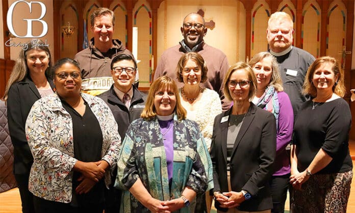 Group photo of eleven diverse adults smiling in a church hall with wooden arches and a 'Giving Back' logo in the corner.