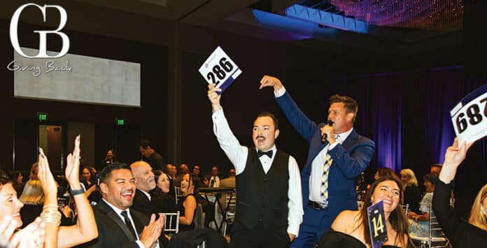 Auction moment at a charity gala: a man holding up a bid paddle while another speaker bids on stage; logo 'GB Giving Back' visible.