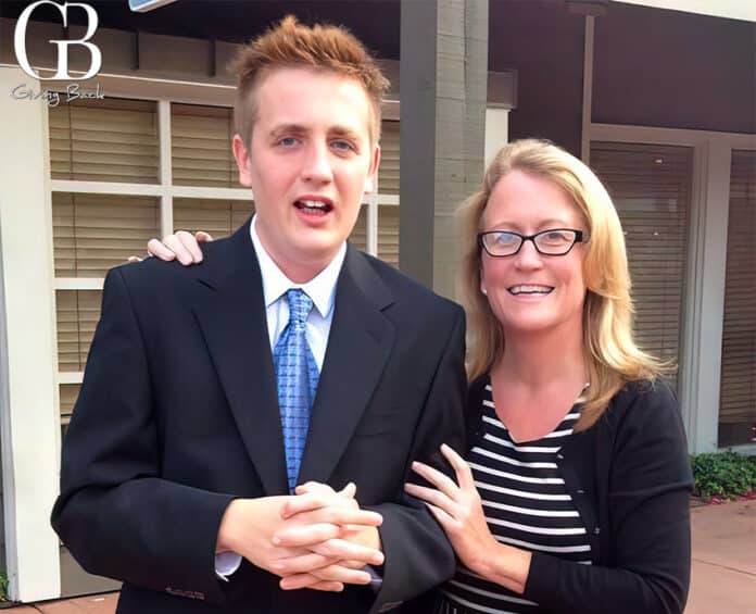 Two people pose for a photo outside a building: a young man in a black suit and blue tie, with a woman in a black cardigan and striped dress smiling beside him. A GB Giving Back logo is in the top-left corner.