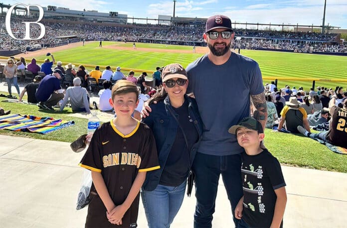 Family of four posing near a baseball field with crowded stands on a sunny day.
