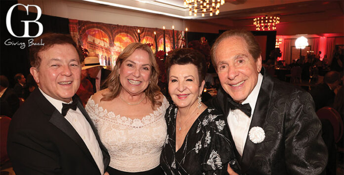 Four adults in tuxedos and evening wear pose together at a charity gala, with the 'Giving Back' logo in the corner of a banquet hall.