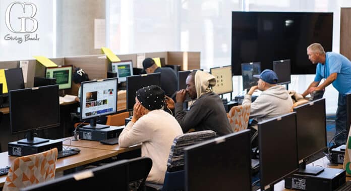 Group of students in a computer lab working at desks with monitors; instructor assists at the back.
