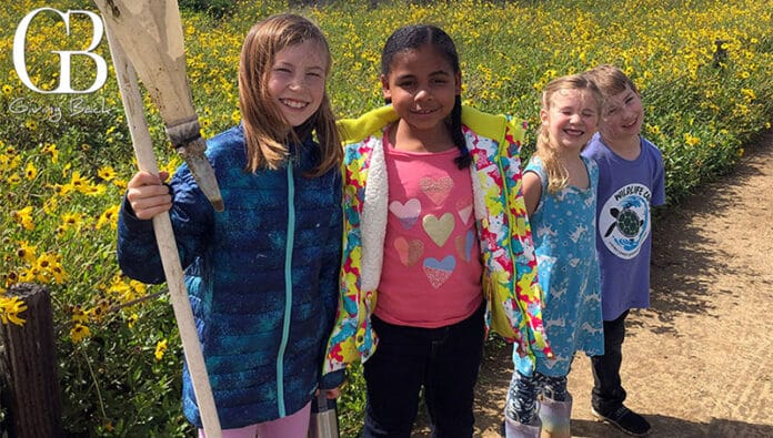 Four smiling children stand on a dirt path in a bright yellow flower field, wearing colorful jackets and shirts.