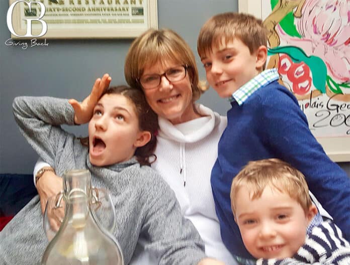 Smiling woman with short hair and glasses sits with three kids posing for a photo indoors.