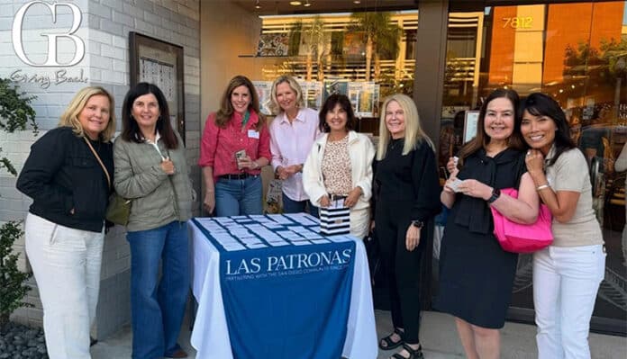 Eight women pose behind a Las Patronas-branded table at an outdoor community event outside a storefront, smiling for the camera.