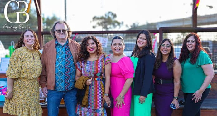 Eight diverse people posing together outdoors at a charity event, smiling for the photo; GB Giving Back logo appears in the top-left corner.