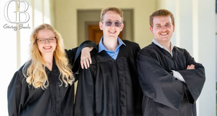 Three graduates in black robes posing on campus, smiling at the camera: a woman with glasses on the left, a man wearing sunglasses in the center, and a man with arms crossed on the right; logo in the top-left corner.