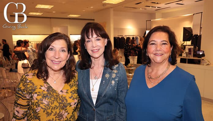Three smiling women pose for a photo at a charity event, with clothing racks and event setup visible in the background and the GB Giving Back logo in the upper left.
