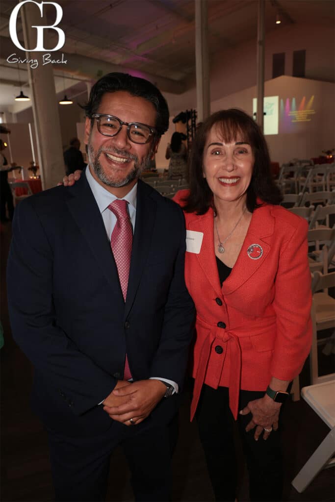 Carlos-Cristiani-and-Julie-Dubick - GB Magazine Carlos Cristiani and Julie Dubick'Giving Back' logo in the upper left corner.","Man in a navy suit and a woman in a coral blazer pose together indoors at a fundraiser, with rows of chairs in the background.","A man with glasses and a woman in a coral blazer stand close together, smiling for the camera at a fundraising event.","Photo of two attendees at a Giving Back event, one wearing a name badge."